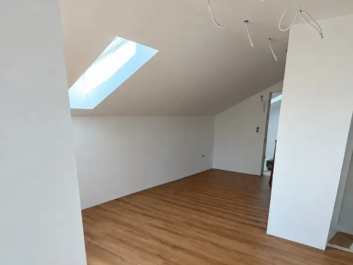 Saniertes Dachgeschoss: weiße Wände, Boden in Holzoptik, Sonnenlicht durch das Dachfenster